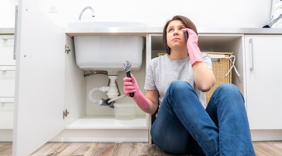 A woman is on the phone as she sits on the floor with a wrench; there is a kitchen sink behind her.