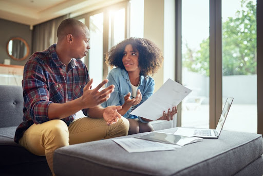 A man and woman sitting on a couch in a home, having a discussion with a laptop and papers scattered in front of them.